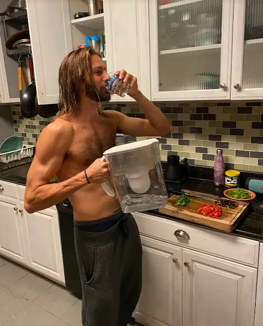a topless guy drinking water from the clearly filtered pitcher in a kitchen